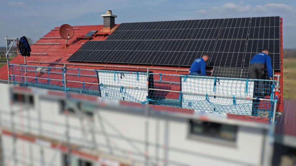Zwei Monteure montieren Photovoltaikmodule auf einem Hausdach, im Vordergrund ist ein Teil der Baustellensicherung zu sehen.