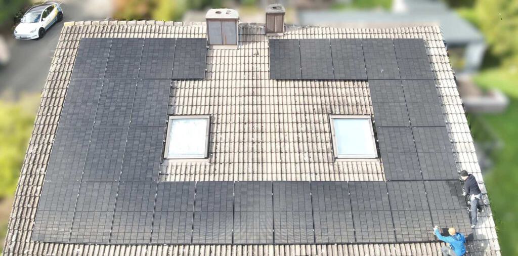 Zwei Monteure installieren schwarze Photovoltaikmodule auf einem Hausdach, aufgenommen aus der Vogelperspektive