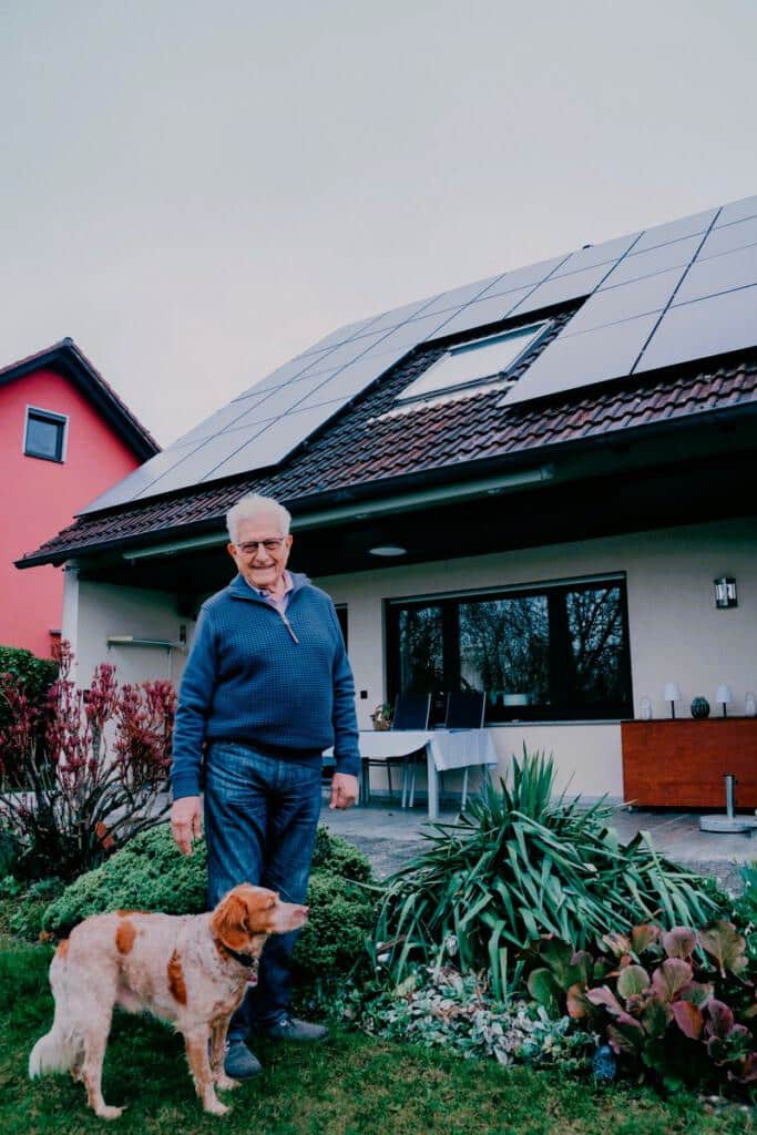 Ein Kunde steht mit seinem Hund im Garten vor einem Wohnhaus, auf dessen Dach Photovoltaikmodule montiert sind.